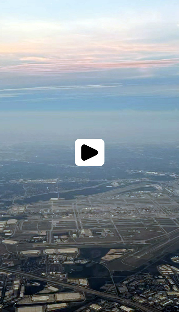 Aerial view of a large airport with multiple runways and terminals, surrounded by buildings and roads, under a cloudy sky during either sunrise or sunset. A play button icon is overlaid in the center. Fearrington Village